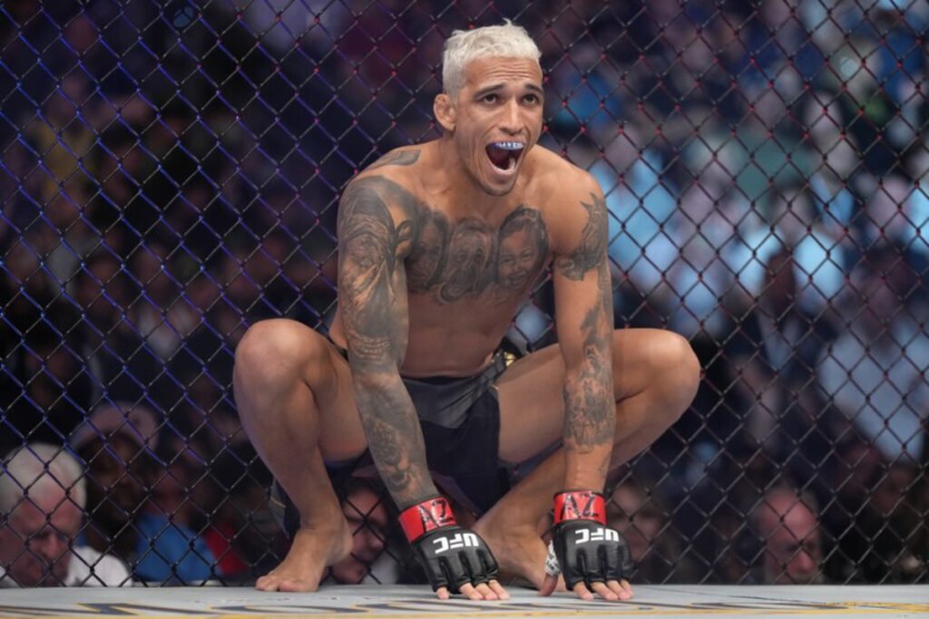 Charles Oliveira, luchador UFC con pelo claro, agachado y gritando en la jaula, con tatuajes y guantes MMA.