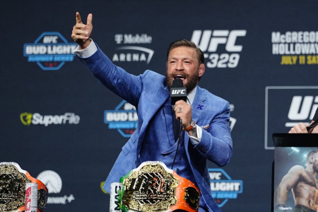 Conor McGregor, radiante en conferencia UFC, con traje, micrófono y cinturones de campeón, alza el brazo triunfante.