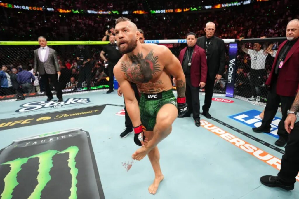 Conor McGregor, en el octágono de UFC, sujeta su pie derecho ensangrentado. Viste pantalón corto verde. Árbitros y público al fondo.