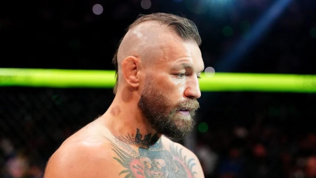 Luchador Conor McGregor de perfil, con barba, cresta y tatuajes en cuello y pecho, en el oscuro octágono de UFC.