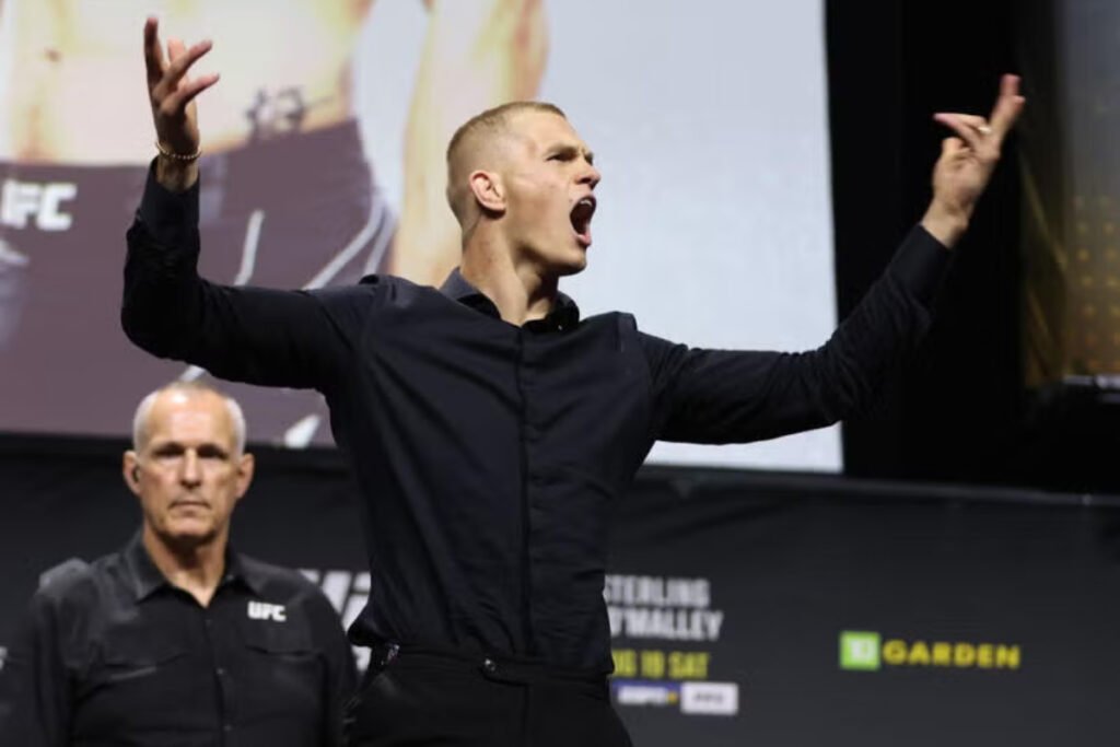 Luchador de UFC, boca abierta y brazos alzados, grita en rueda de prensa. Un oficial observa serio de fondo.