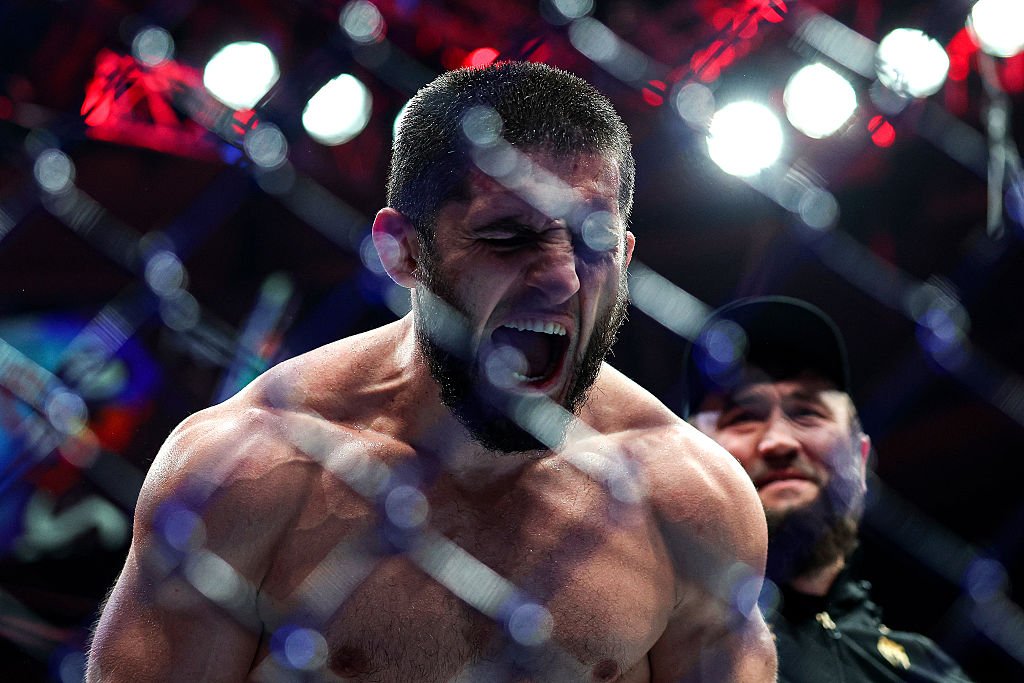 Islam Makhachev ruge eufórico en la jaula de MMA, mostrando su musculatura. Fondo desenfocado con luces y un hombre sonriente.
