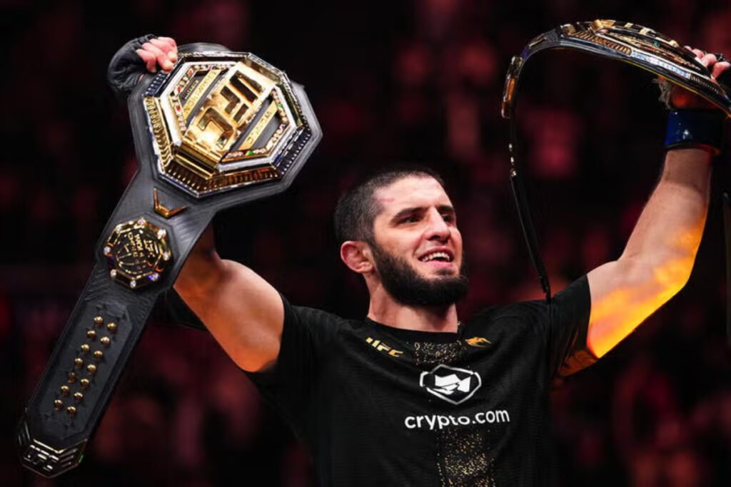 Islam Makhachev, luchador de MMA, sonríe triunfante levantando sus dos cinturones de campeón de UFC.