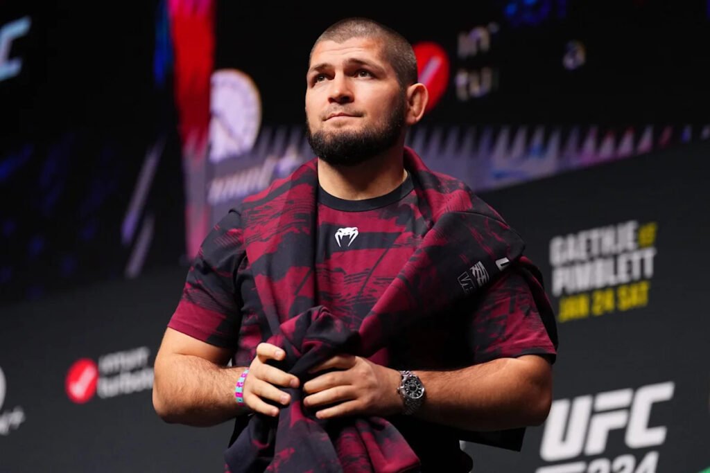 Khabib Nurmagomedov con barba y camiseta Venum roja y negra, en un evento UFC. Mira pensativo hacia arriba.
