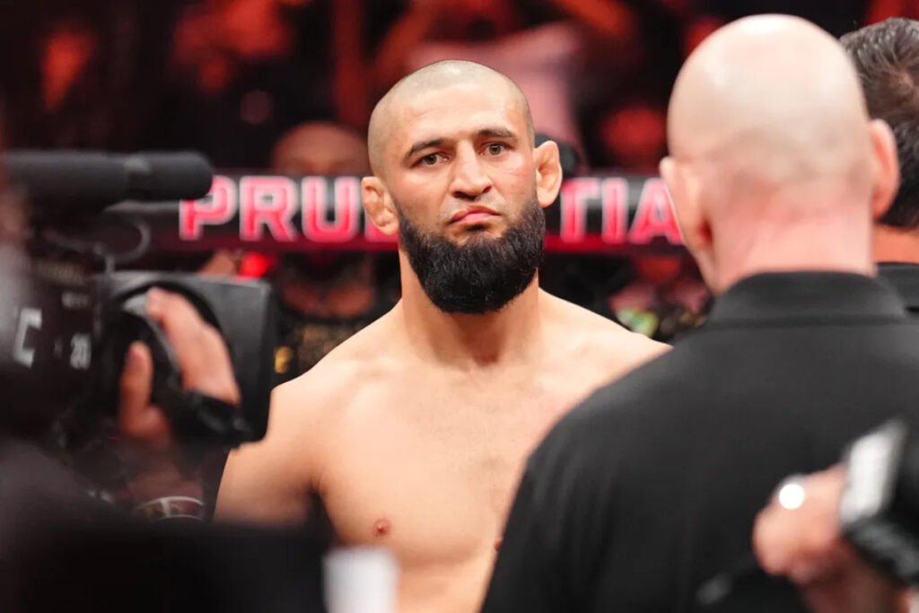 Khamzat Chimaev, luchador de UFC con barba y cabeza rapada, mira fijo. Un árbitro le da la espalda. Luces rojas de fondo pre-combate.