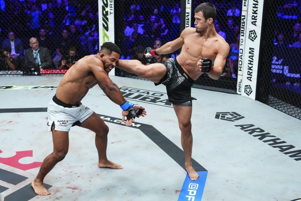 Lucha MMA: Un peleador lanza una patada potente a la cara de su oponente, que reacciona al impacto en la jaula. Combate intenso.