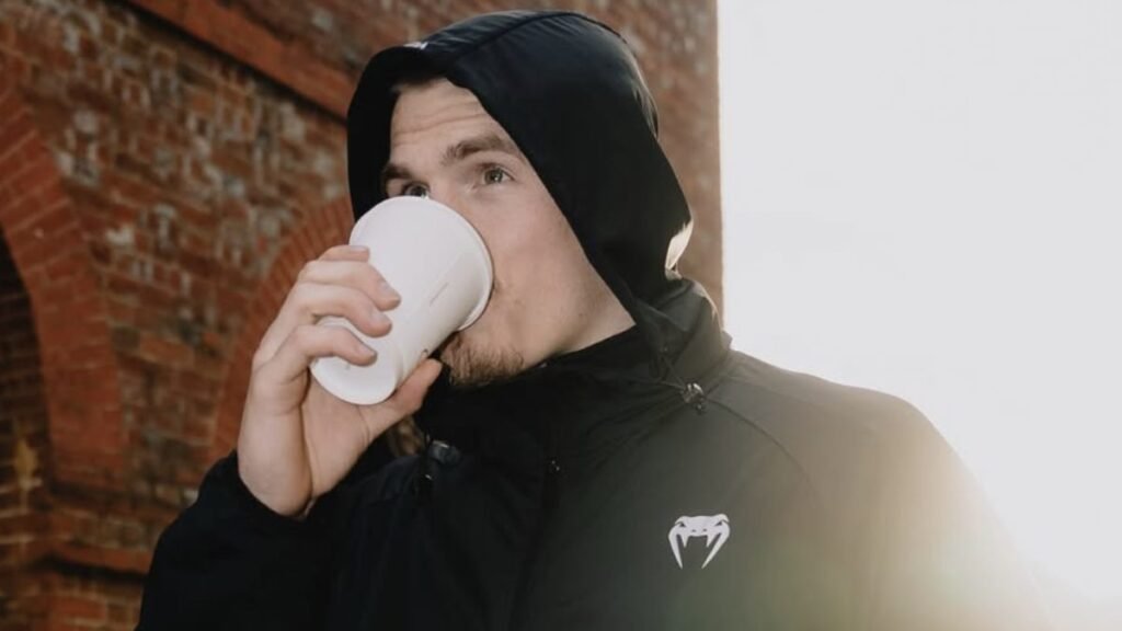 Hombre con sudadera negra Venum bebe de un vaso blanco al aire libre, con luz solar. Barba y mirada atenta.