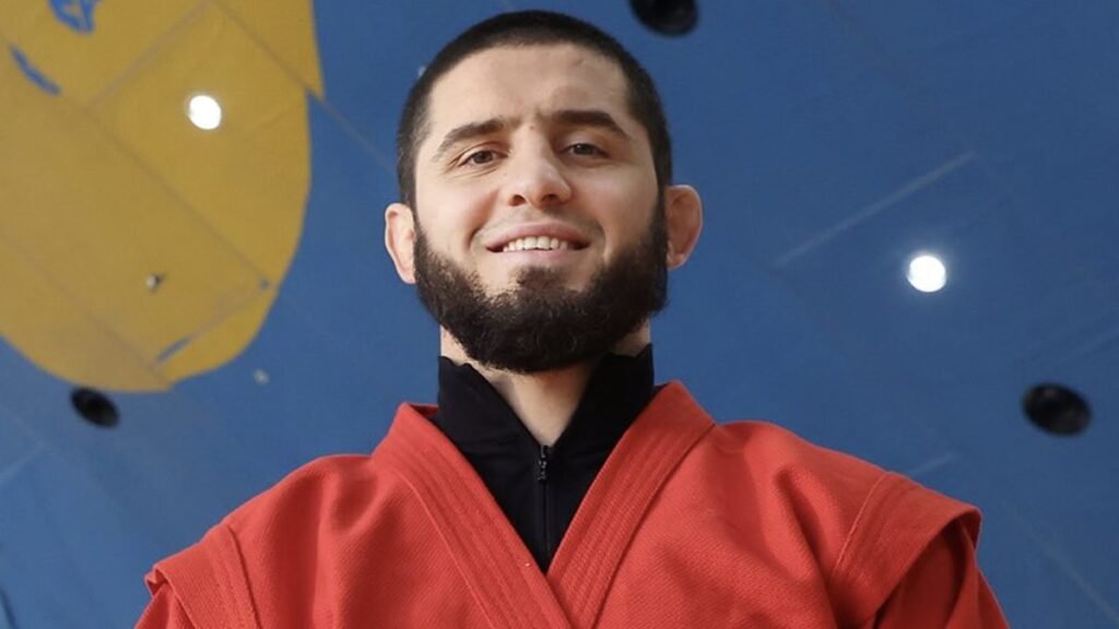 Hombre sonriente con barba, vistiendo un uniforme rojo de Sambo sobre chaqueta negra. Fondo azul con paneles amarillos y luces.