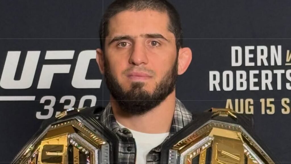 Islam Makhachev, campeón de UFC, con su cinturón de oro y negro, mirando a cámara. Fondo de cartel UFC 330.