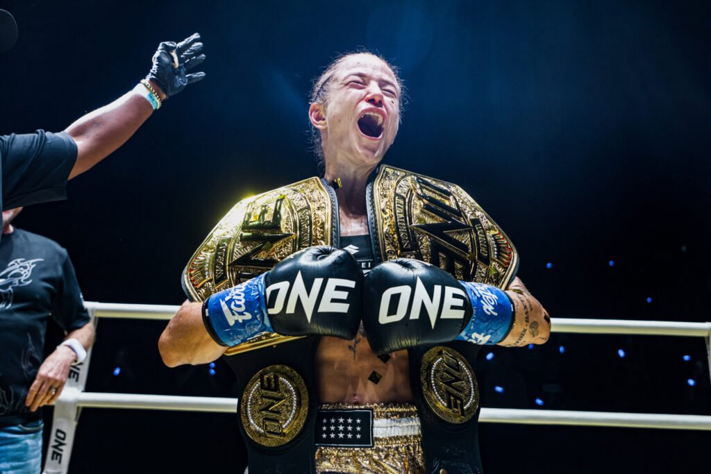 Boxeadora campeona gritando de victoria, luciendo cinturón dorado y guantes ONE en el ring.