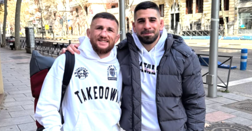 Dos luchadores barbudos y sonrientes, con orejas de coliflor, posando en una calle urbana. Uno lleva sudadera Takedown.