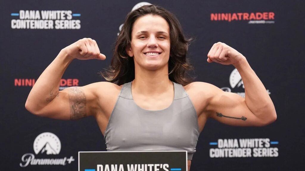 Luchadora de MMA sonriente, flexiona bíceps con tatuajes. Top gris. Fondo de Dana White's Contender Series.