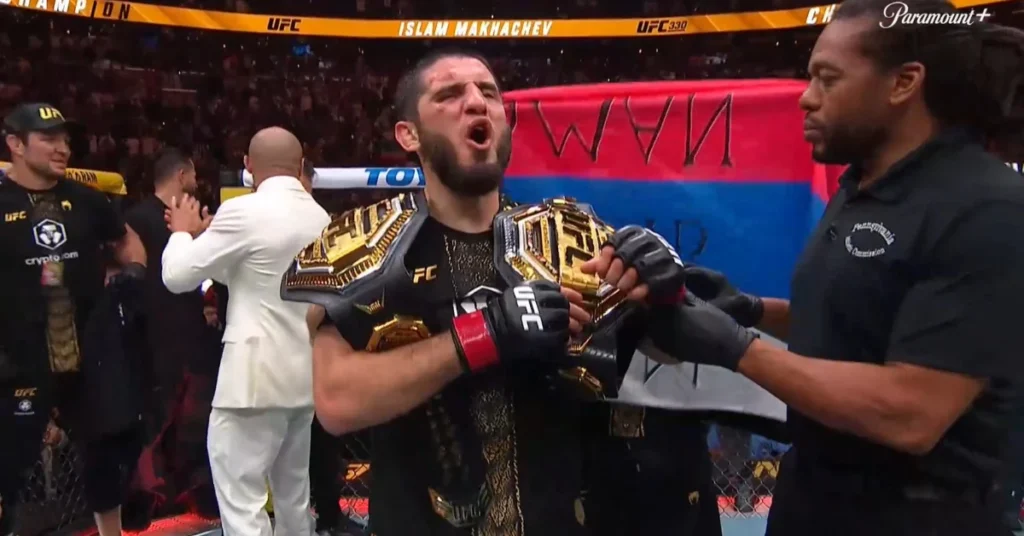 Islam Makhachev, campeón UFC 330, gritando de júbilo con dos cinturones en el ring tras su victoria.
