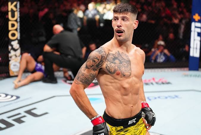 Luchador de UFC con tupé, bigote y tatuajes extensos, guantes rojos y negros, mira con la boca abierta en el octágono.