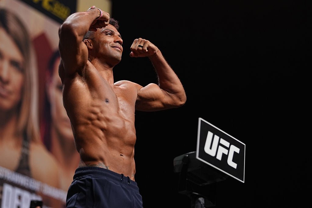 Atleta musculoso de UFC posa sonriente, mostrando bíceps y abdominales definidos en el escenario.