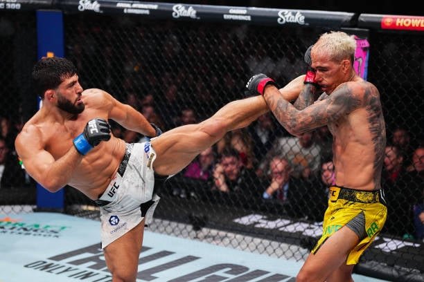 Luchador con barba y shorts blancos lanza patada alta a oponente tatuado de pelo rubio en shorts amarillos bloqueando en UFC.