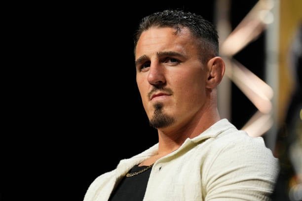 Hombre con oreja de coliflor y barba de chivo, mirada intensa. Lleva camisa crema, cadena de oro. Un luchador de MMA en una conferencia.