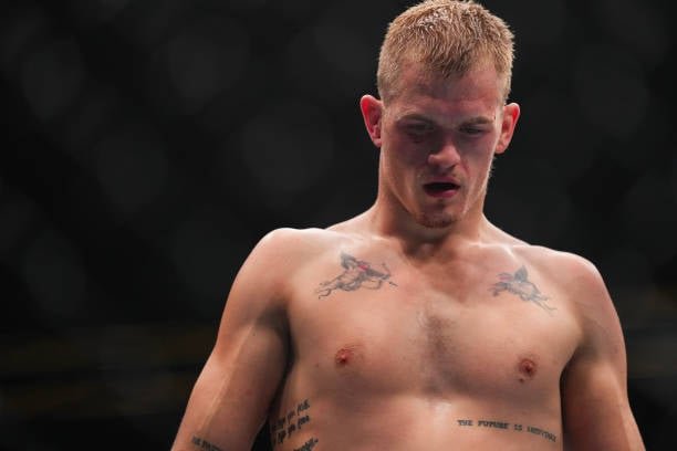 Luchador de MMA rubio, sin camiseta, con tatuajes en el pecho, mira hacia abajo, exhausto.