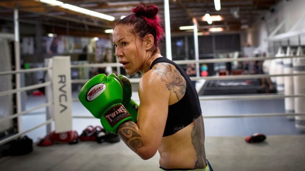 Mujer con pelo rojo y guantes de boxeo verdes Twins Special en un ring. Tatuajes visibles, sudando, mira intensamente durante el entrenamiento.