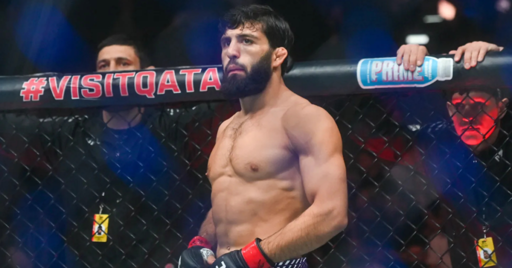 Luchador de MMA con barba y torso desnudo en el octágono. Lleva guantes y patrocinios #VisitQatar y Prime en la valla.