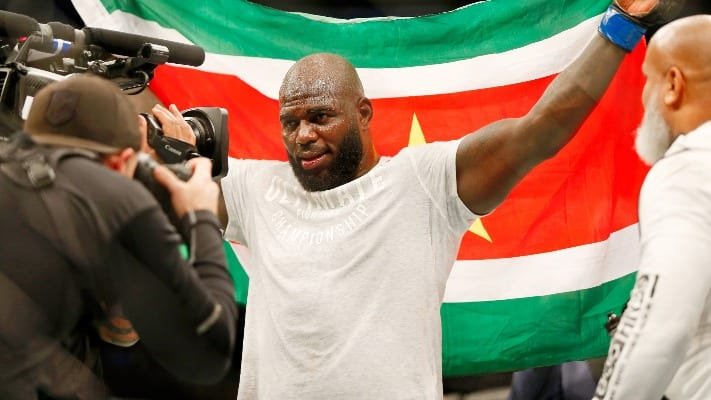 Luchador calvo y barbudo, sudoroso, celebra con la bandera de Surinam mientras un cámara y su entrenador le observan.