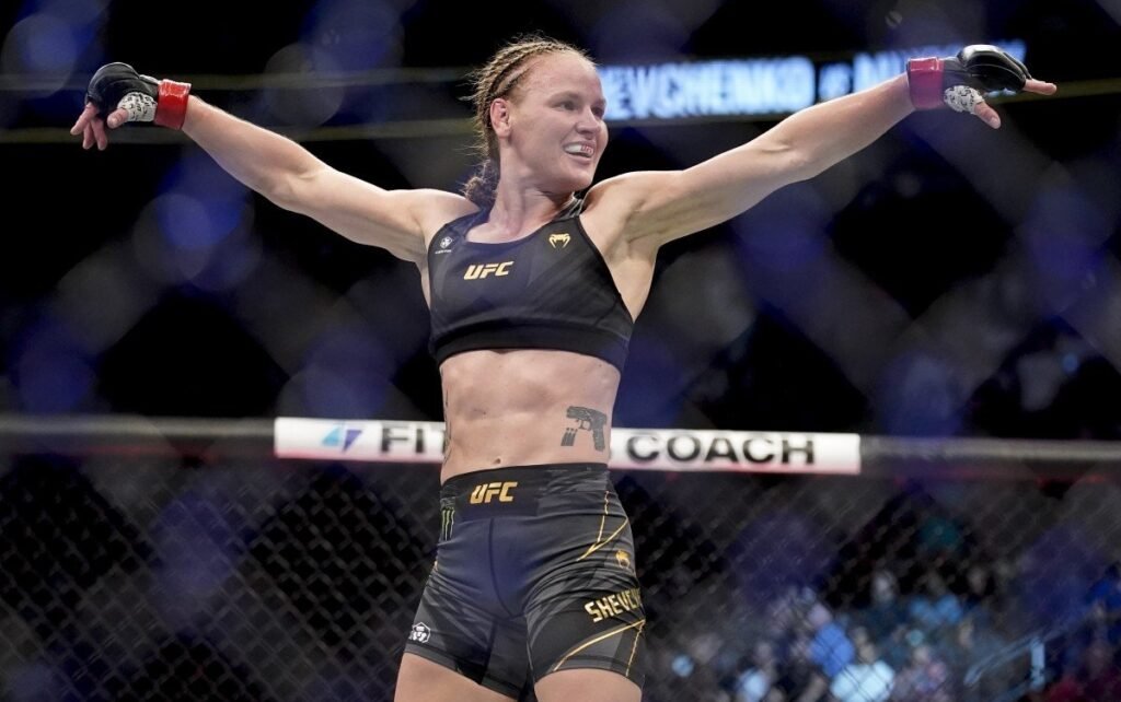 Valentina Shevchenko de UFC celebra victoria en la jaula, sonriendo con brazos en alto. Destaca su tatuaje de pistola.