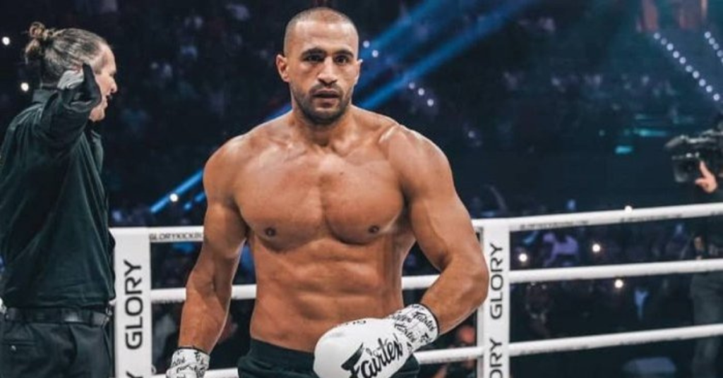 Peleador de GLORY, musculoso y sin camiseta, en el ring con guantes Fairtex, mostrando concentración. Un árbitro le observa.