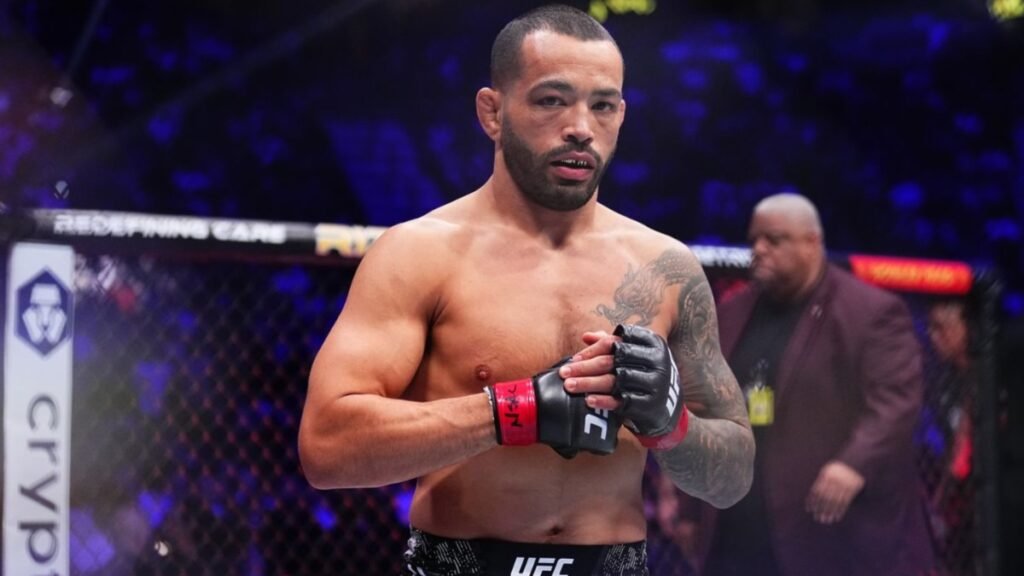 Dan Ige, luchador de UFC, con tatuajes y protector dental dorado, mira a cámara dentro de la jaula. Público difuso.