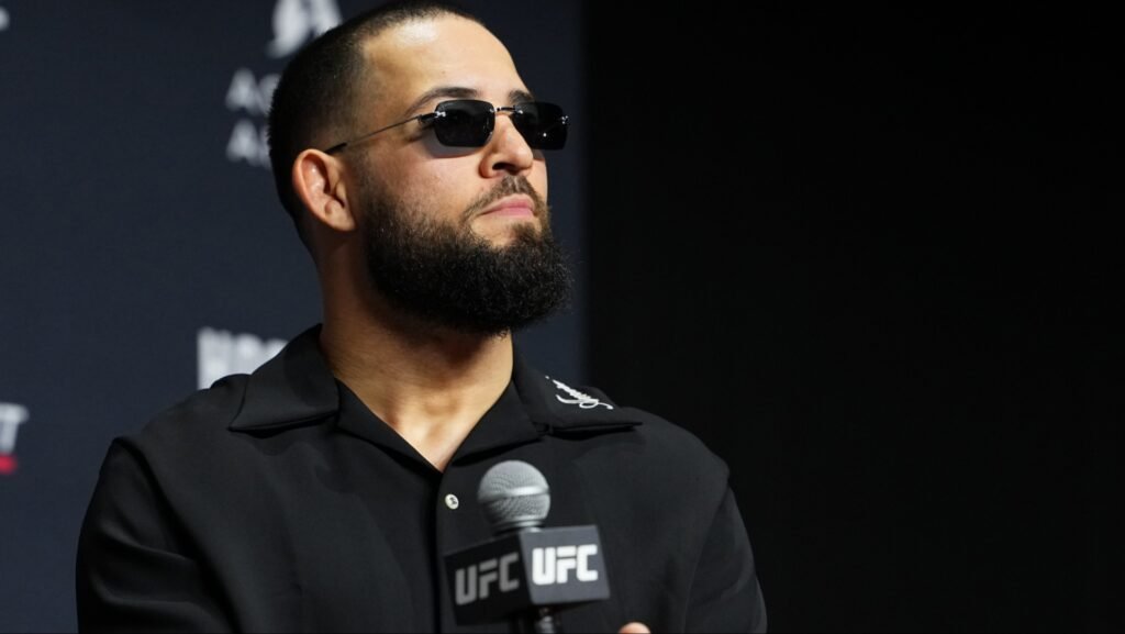 Hombre barbudo con gafas de sol y camisa polo negra. Sostiene un micrófono UFC, con mirada pensativa en un evento.