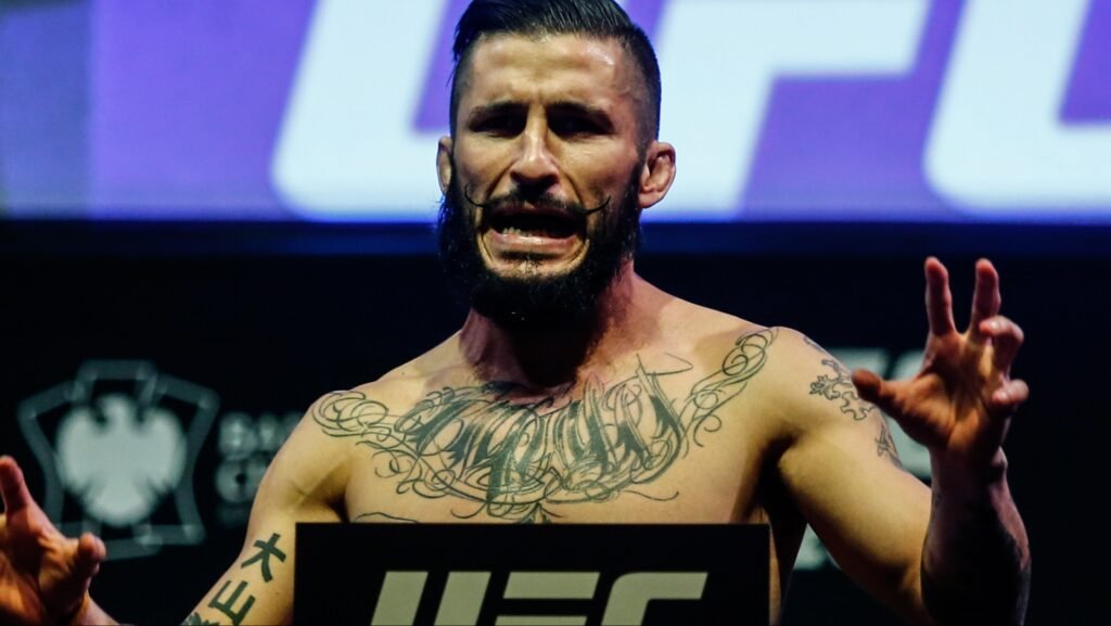 Luchador de MMA con tatuajes y bigote, expresión intensa en pesaje UFC. Manos alzadas, fondo púrpura.