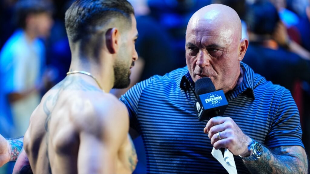Joe Rogan (calvo, camisa a rayas) entrevista a un luchador de la UFC sin camiseta y tatuado. Sujeta un micrófono de UFC Freedom 250.