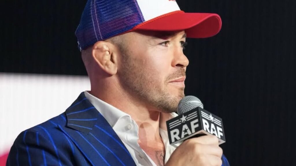 Colby Covington, luchador UFC, de perfil con gorra tricolor, traje azul de rayas y micrófono RAF. Oreja de coliflor visible.