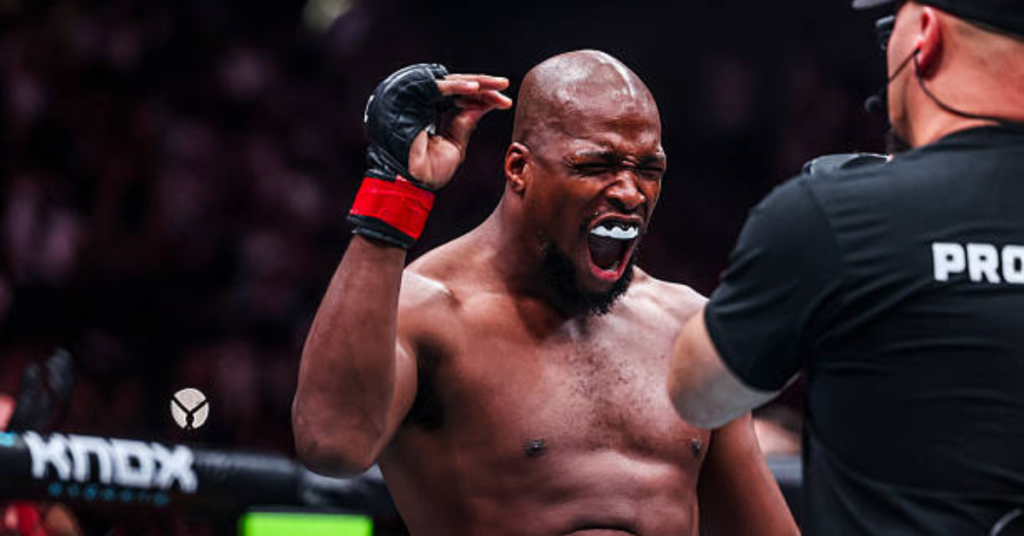 Luchador de MMA negro celebra con euforia en el ring, boca abierta con bucal y puño en alto. Árbitro de combate al lado.