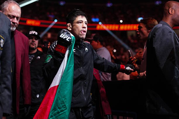 Brandon Moreno, luchador UFC, con la bandera de México y guante de combate, camina en una arena. Fondo con espectadores.