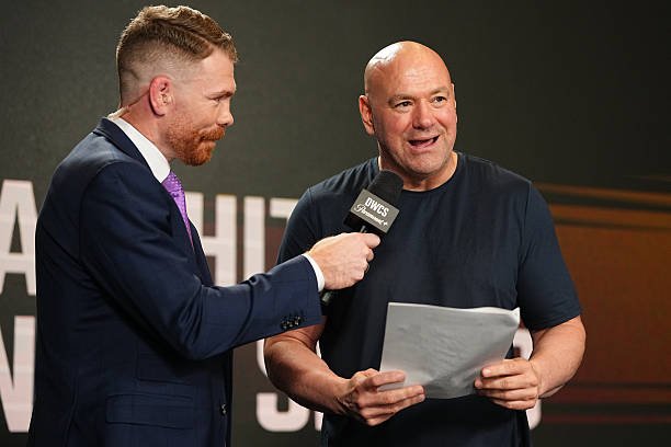 Presentador con barba y traje entrevista a Dana White, presidente de la UFC, quien sonríe sujetando documentos con micrófono.