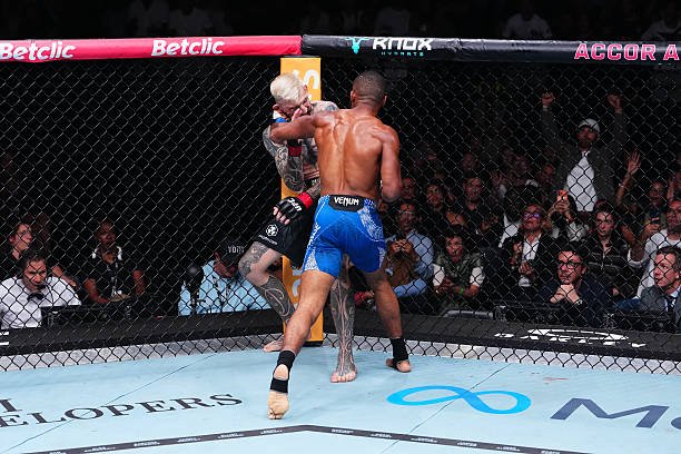 Combate de MMA en octágono: un luchador rubio tatuado recibe un puñetazo de un oponente con shorts azules. Público animado.